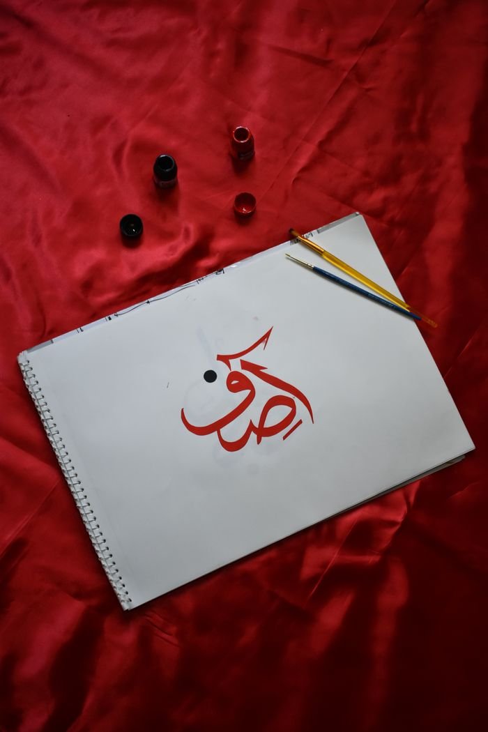 Creative Arabic calligraphy in red ink displayed on a notebook with paintbrushes.