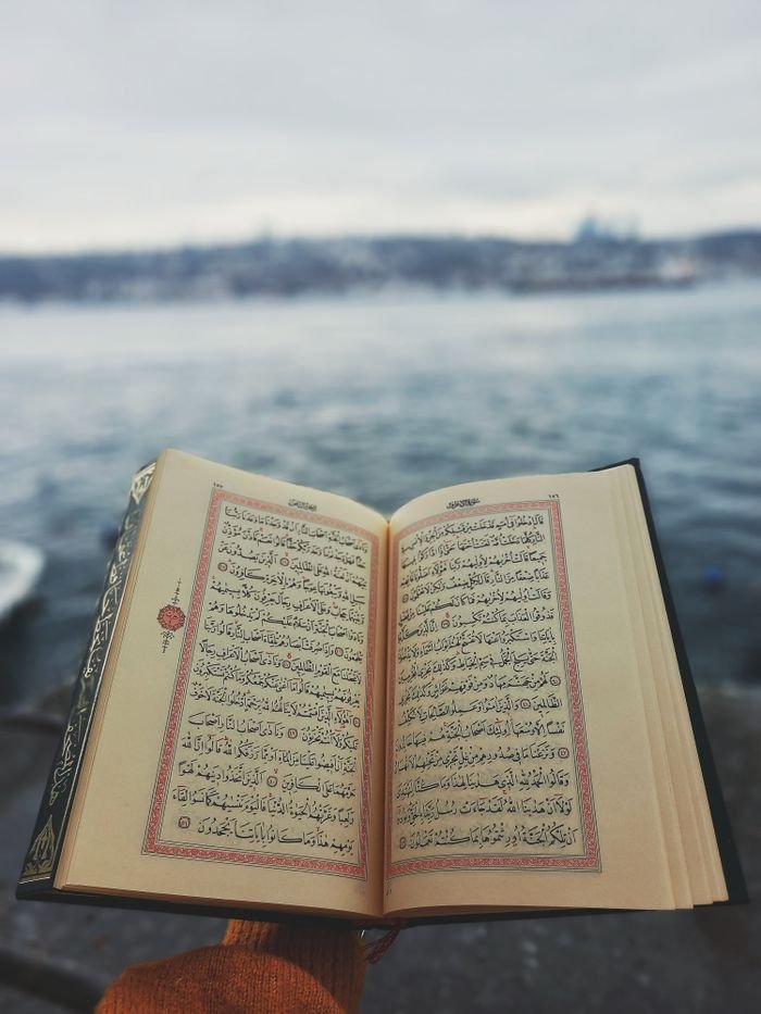 Open Quran being held on a riverbank with city skyline in the background.