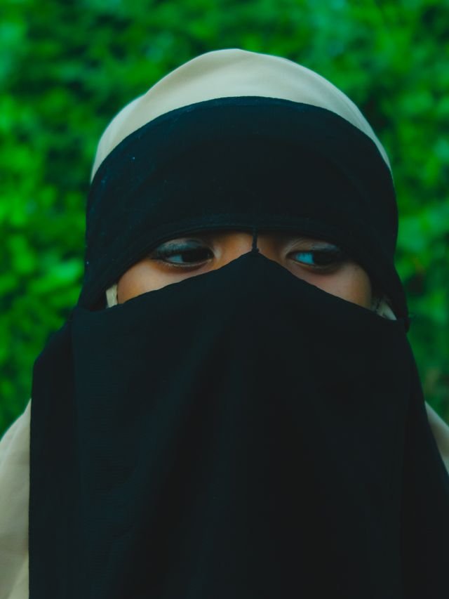 A close-up portrait of a woman wearing a black niqab, set against a green leafy background.