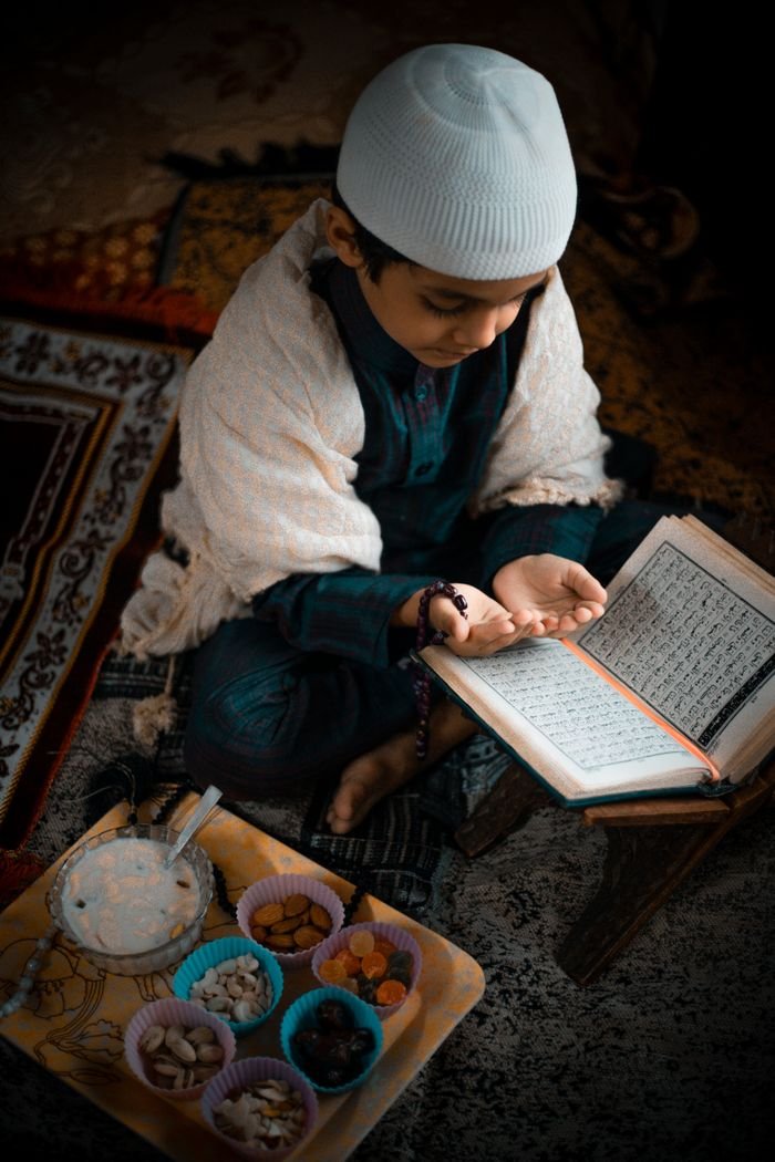 A young boy in traditional attire reads and prays with an open Quran beside snacks.
