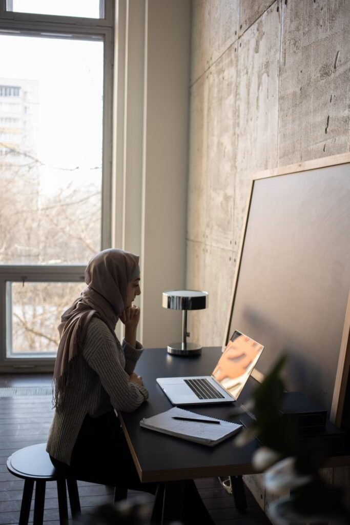 Side view of serious Muslim female student in headscarf browsing netbook at table with copybook and chalkboard while doing task for lesson